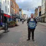 Alex McIntyre MP standing on Westgate Street holding a Shine a Light on Local bauble.